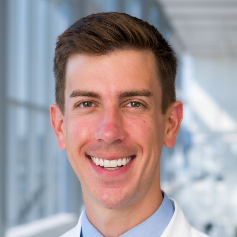 Smiling young man with brown hair wearing a blue dress shirt, dark blue tie, and a white lab coat.